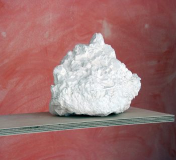 Detail of a birch plywood shelf on a freestanding plywood sheet with a plaster cast rock against a washed out terracotta coloured wall.