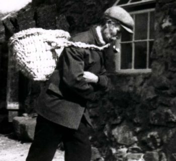 a film still in black and white, a man walking hunched forward, pipe in mouth and a cap, arms crossed carrying a weaved basket on his back