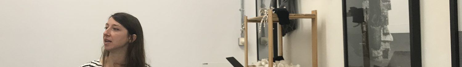 Oana Stanciu in studio during studio visit, gesturing, with objects and prints on the wall.