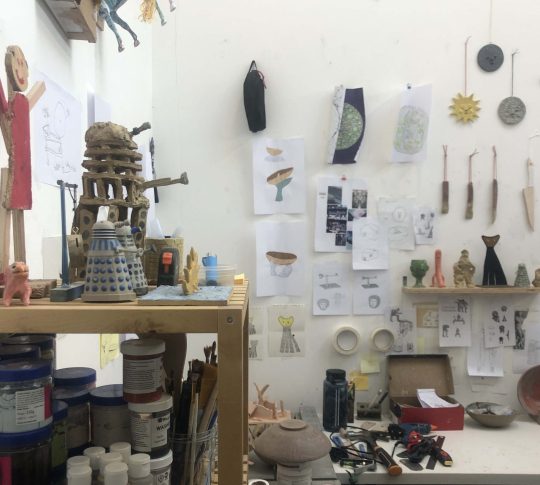 Studio photo shelves with materials and items, and drawings on the facing wall above a desk